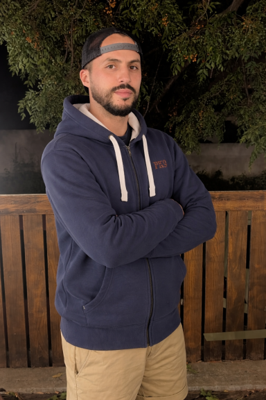 Homme avec un sweat à capuche bleu, bras croisés devant une clôture en bois.